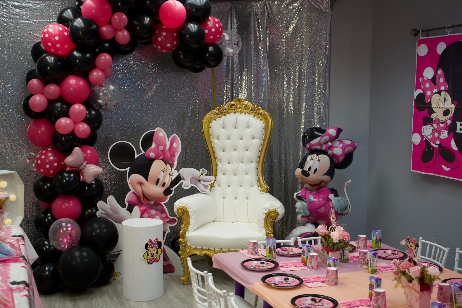 Minnie Mouse themed birthday party room with white and gold throne chair, pink and black balloon arch, Minnie Mouse decorations, pink table settings and flowers at Star Park